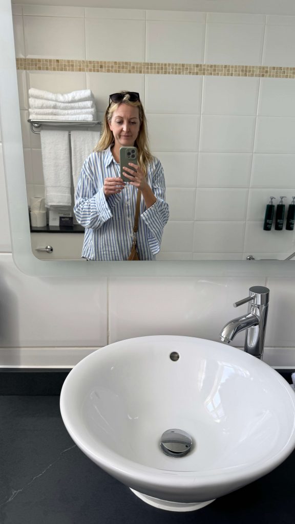 Woman taking a mirror selfie in a bathroom with a sink and towels.